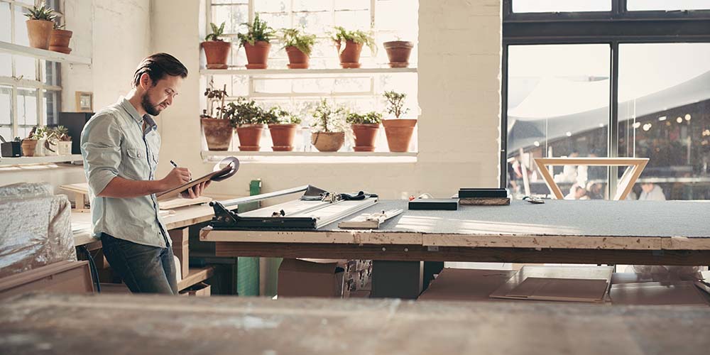 Young male business owner standing in his studio workshop checking figures and stock on his clipboard on a sunny afternoon